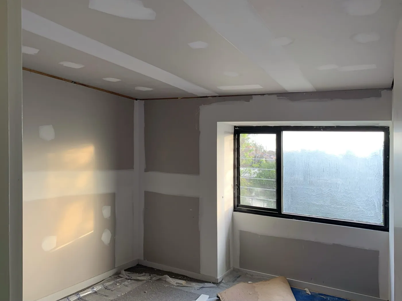 Bedroom before, plasterboard with set joints and patches around a black framed window, Caulfield East